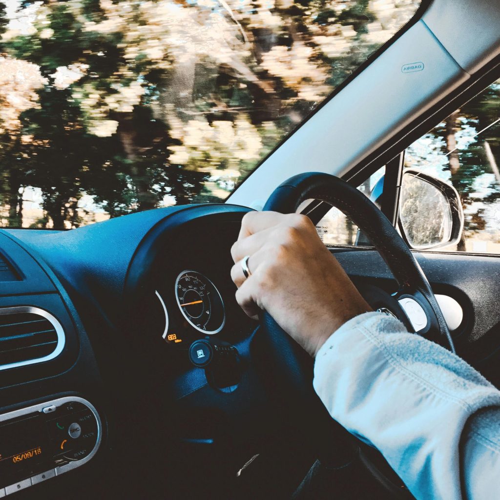 Driver's hand on steering wheel of a modern car during a daytime drive through scenic outdoors.