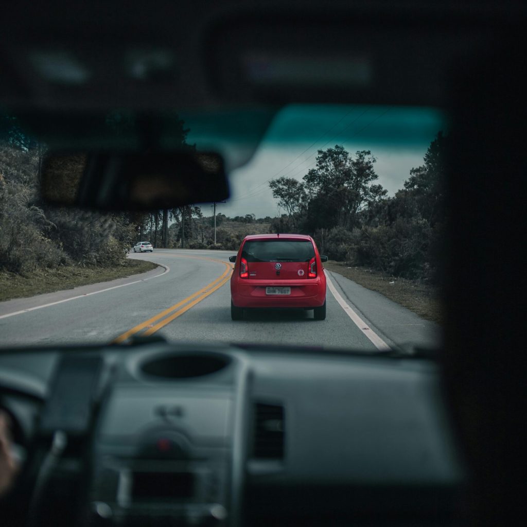 A red car driving on a winding countryside road, viewed from inside another vehicle.